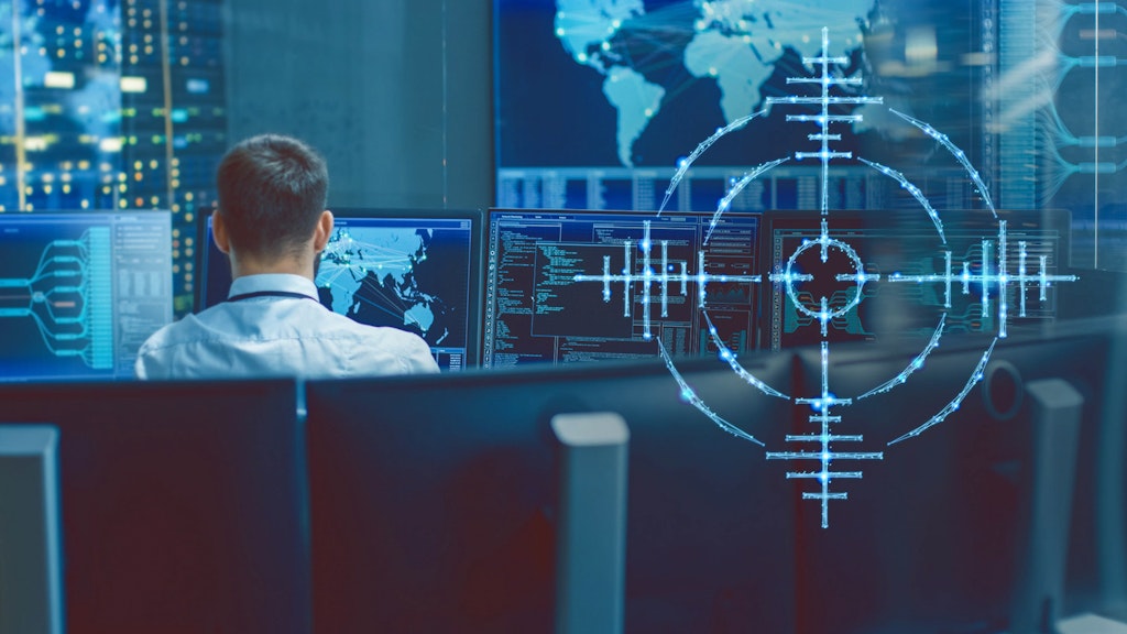 A target cross can be seen in the foreground. In the background a person is sitting at computers.