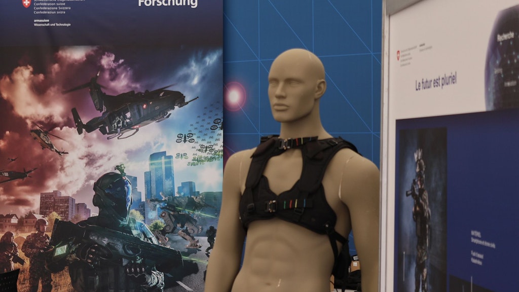 We can see part of the armasuisse S+T exhibition area: Posters, a doll, a table with brochures and a screen from behind. 