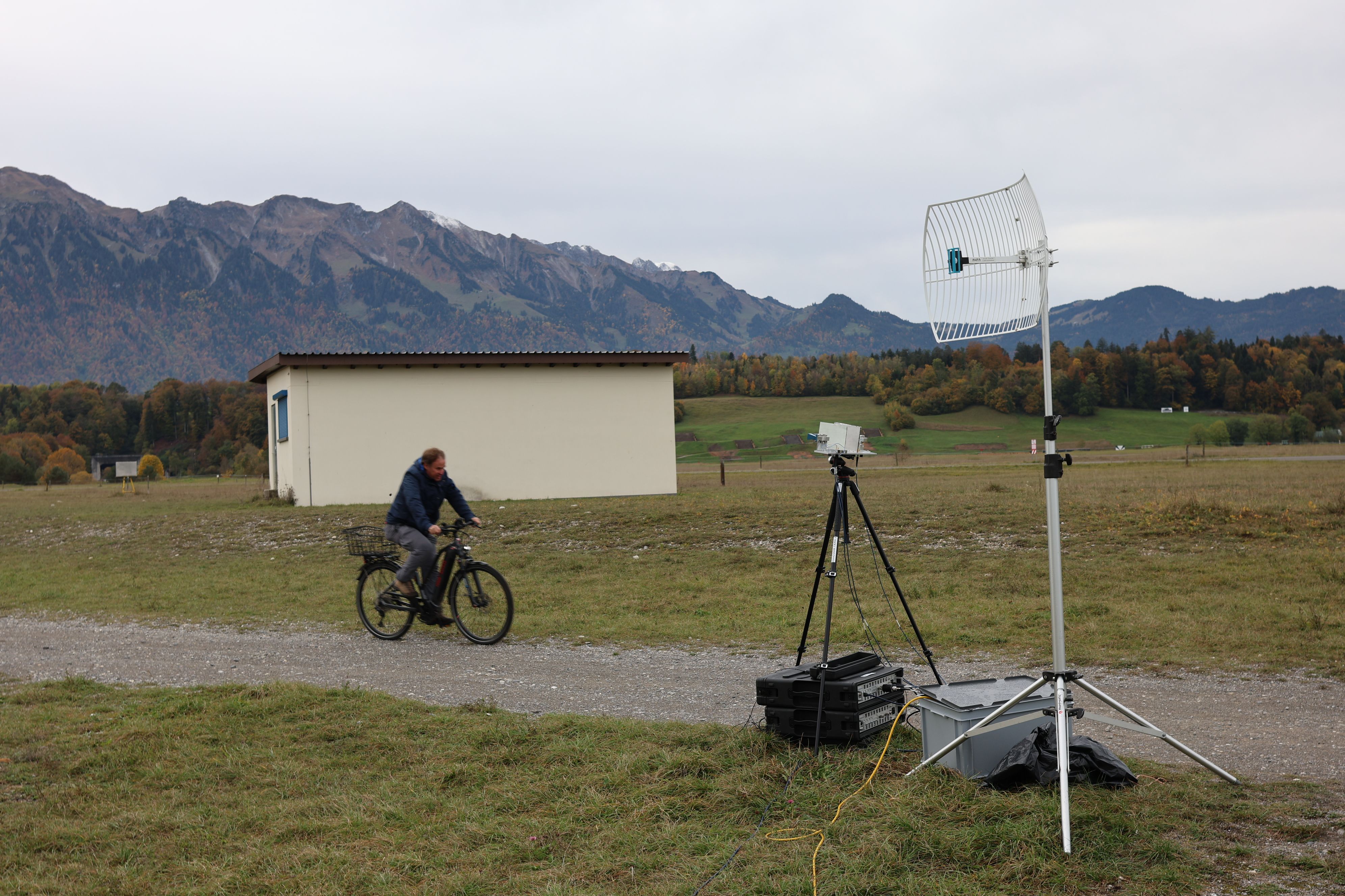 In the foreground, we can see two boxes, an antenna and a black tripod. A man approaches them on a bicycle, with a small house and nature in the background. 