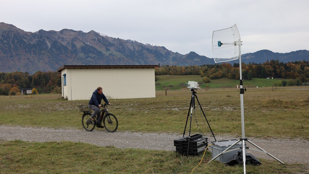 In the foreground, we can see two boxes, an antenna and a black tripod. A man approaches them on a bicycle, with a small house and nature in the background. 