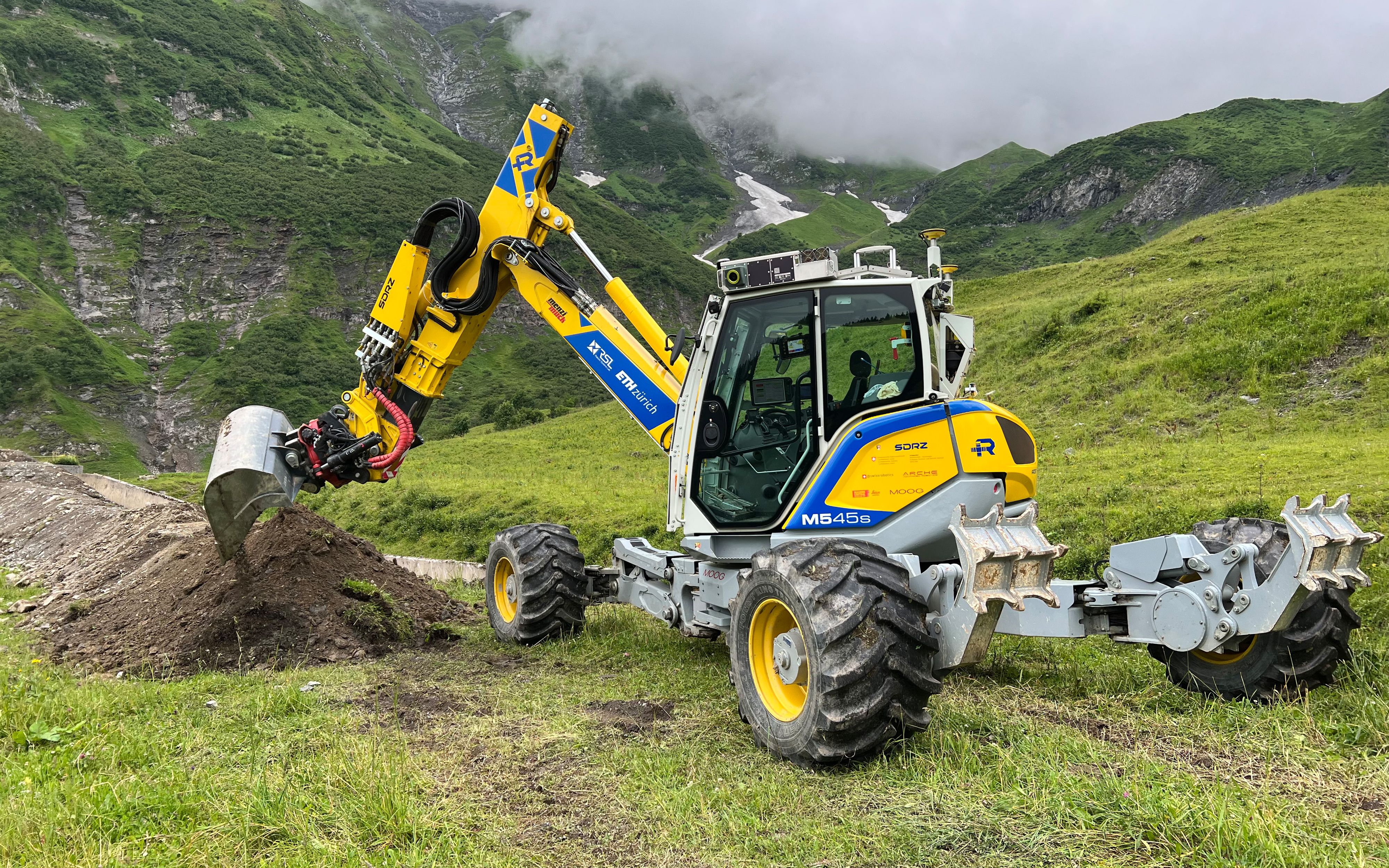 An unmanned, yellow excavator, which is operated by remote control and is in the process of dig-ging a trench. Nature can be seen in the background. 