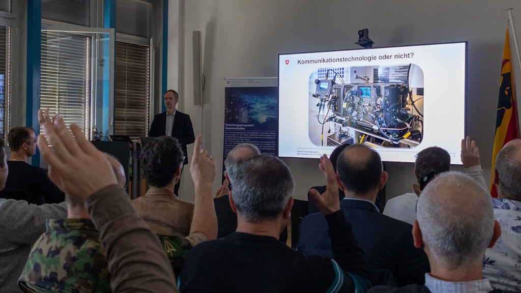 A man stands at a podium and presents a PowerPoint slide on a large screen. Facing him sits an audience, some of whom are raising their hands. 