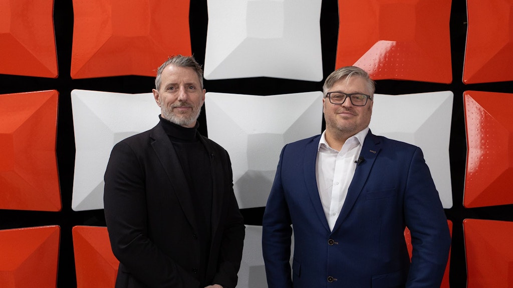Two men in suits stand in front of a large Swiss cross. 
