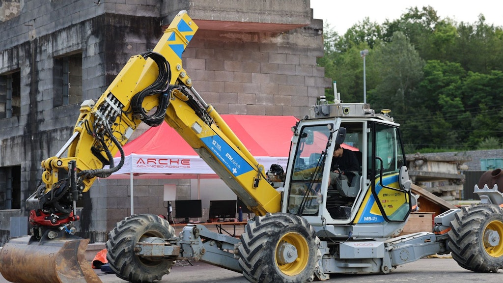 A man is sitting in a yellow walking excavator and behind the excavator is a red tent with the lettering ARCHE.