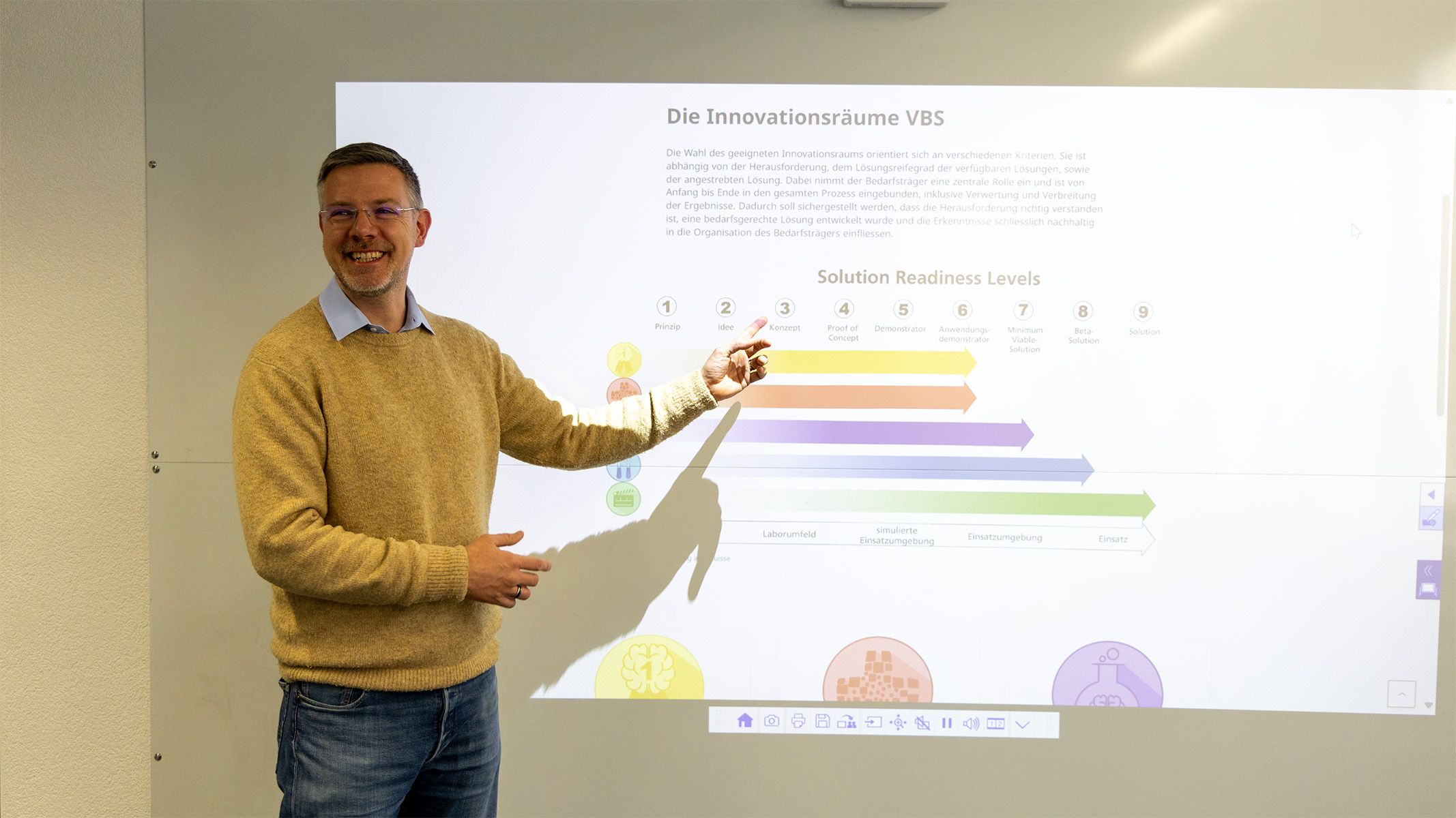 Dr. Jens Rehanek is standing in front of a white wall. He points to the presentation that is projected.