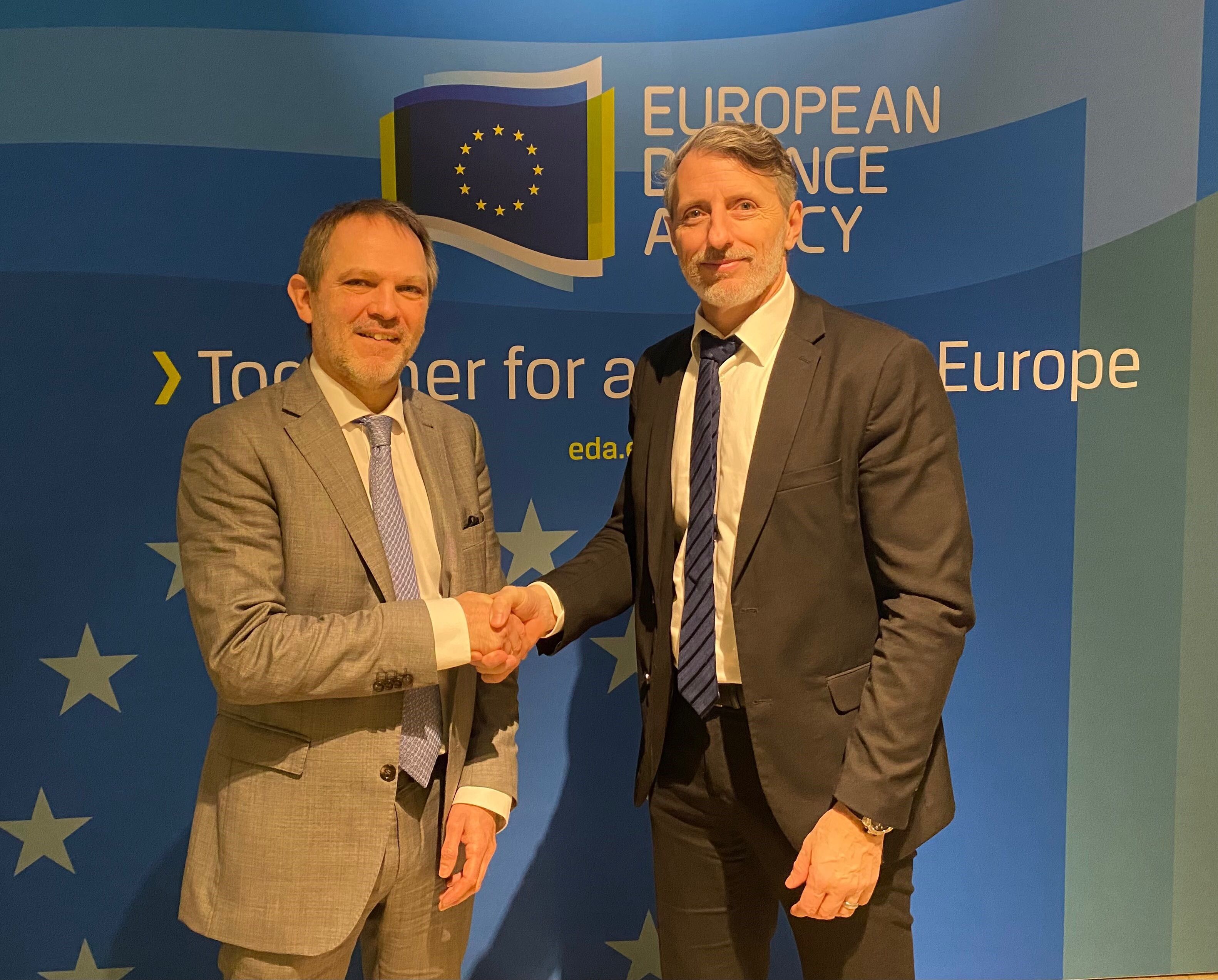 Jean-Francois Ripoche, links im Bild, und Thomas Rothacher, rechts im Bild, stehen vor einer EDA-Tafel, schütteln sich die Hand und schauen dabei in die Kamera.