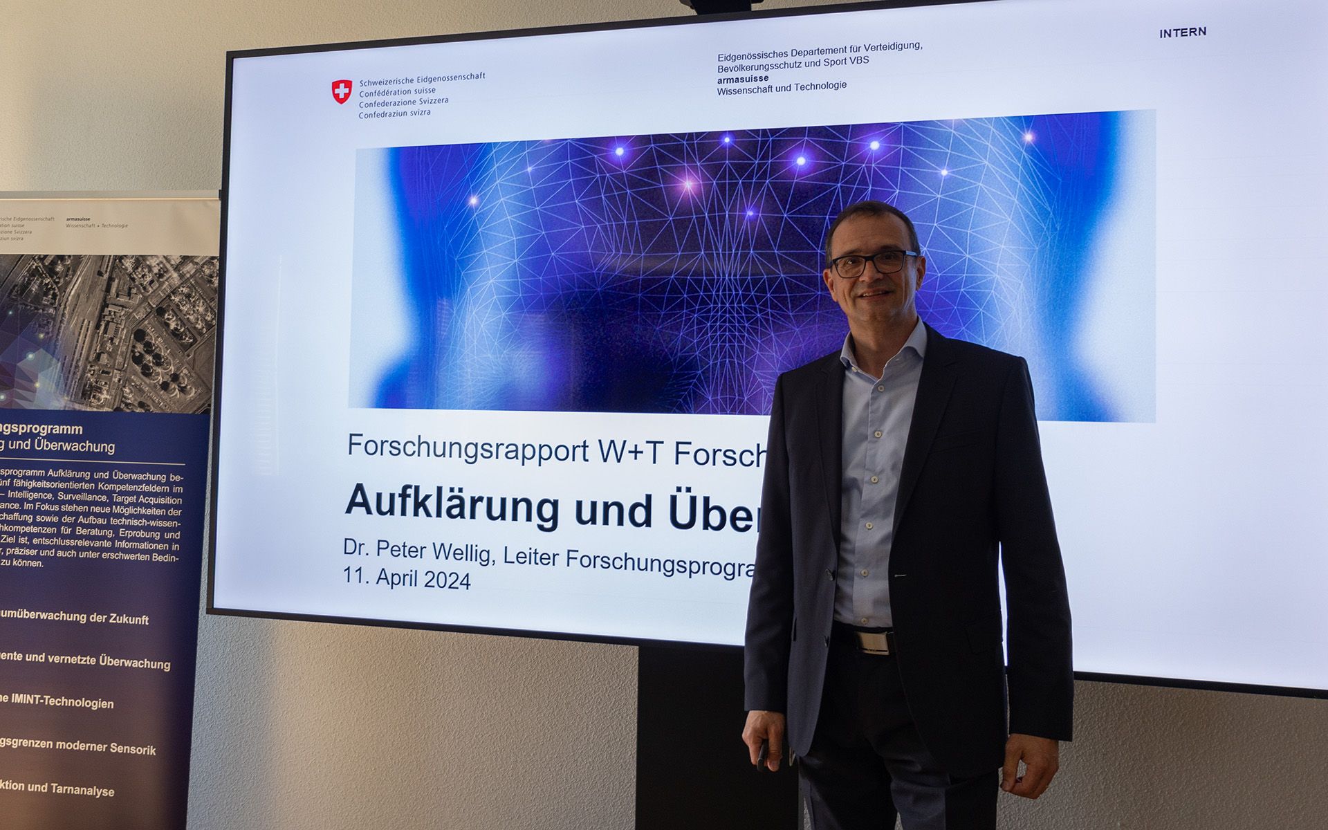 Ein Mann steht vor einer Leinwand, auf welcher eine Präsentation zu sehen ist.