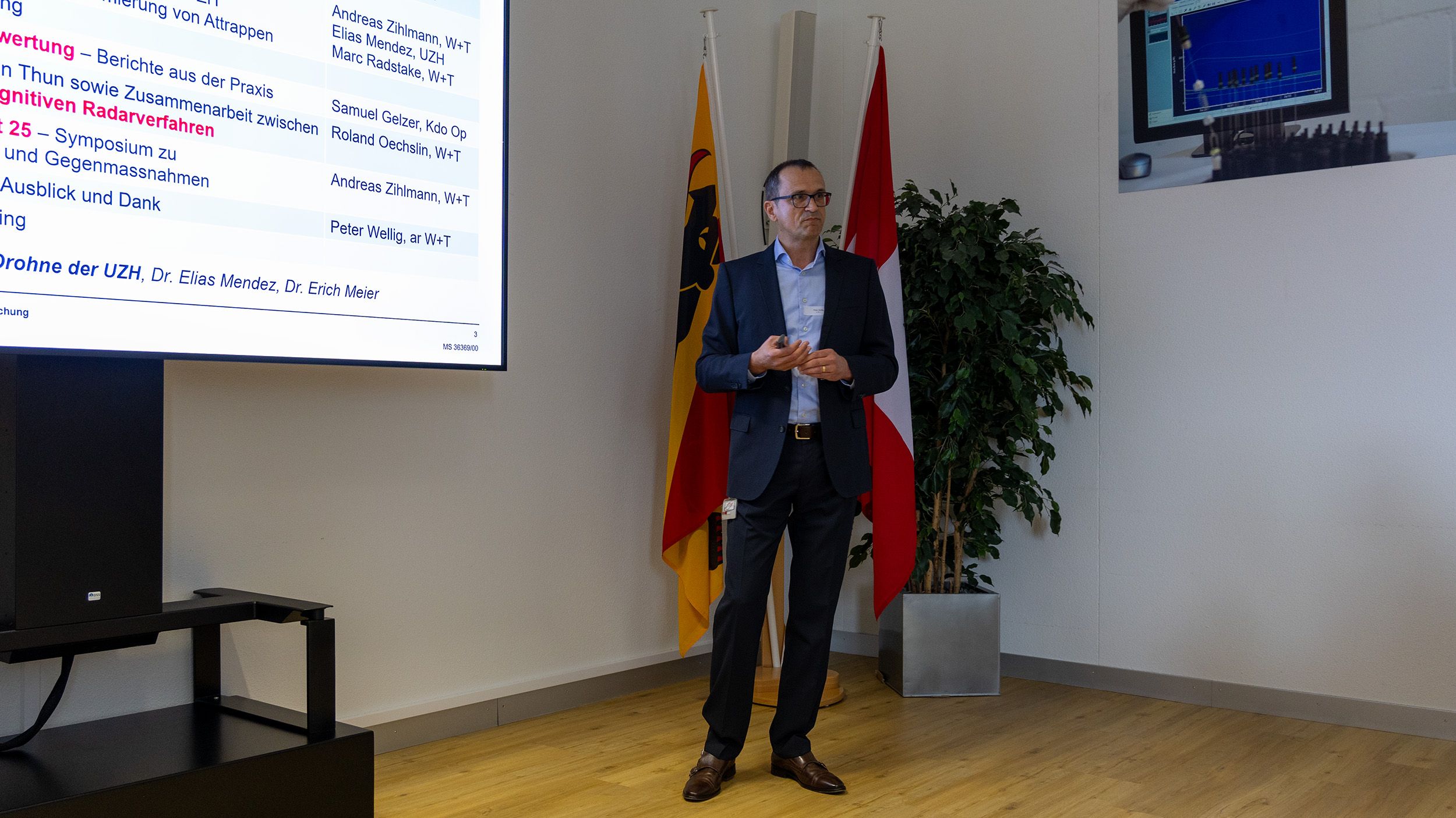 Dr. Peter Wellig steht in der Ecke eines Raumes neben einem Bildschirm, auf dem eine Präsentation zu sehen ist. Hinter ihm befinden sich zwei Fahnen.