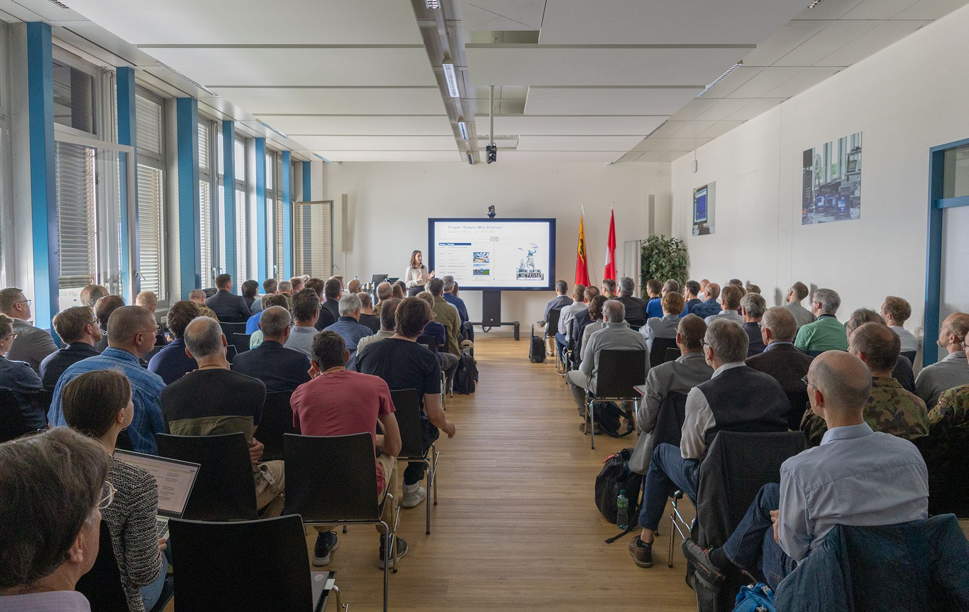 Eine Frau präsentiert vor einem Publikum in einem grossen Raum. Neben ihr befindet sich ein Bildschirm. 