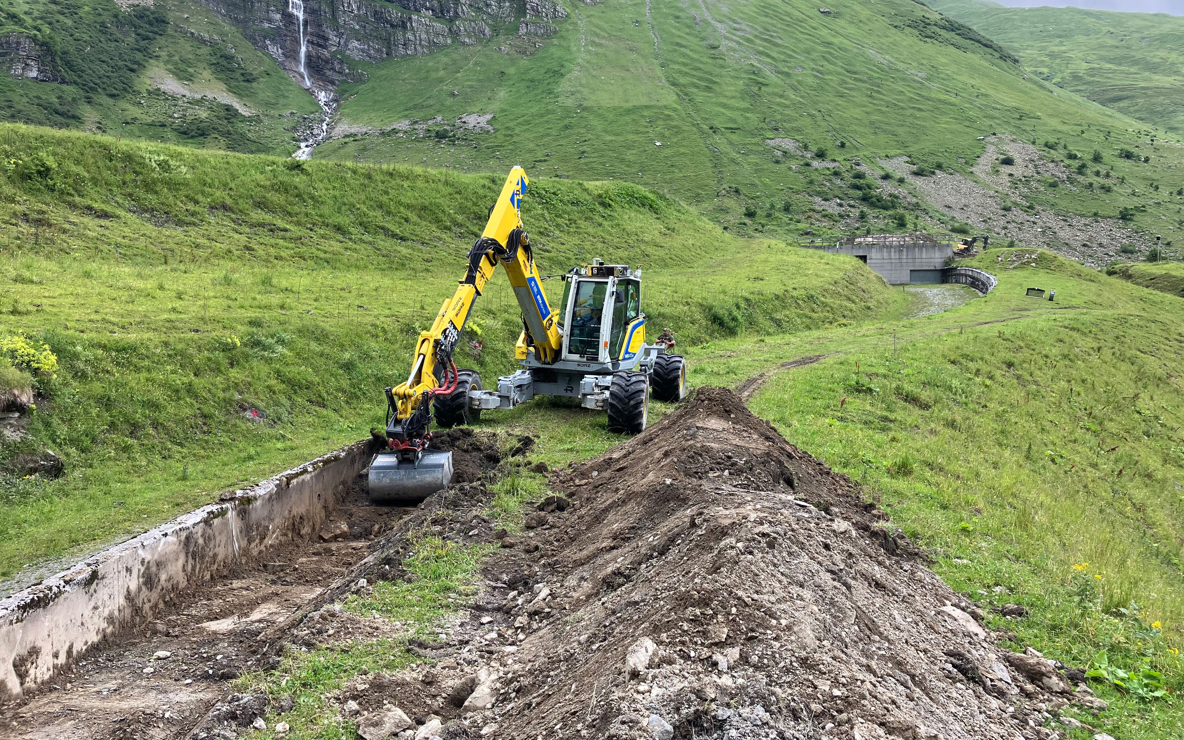 Ein unbemannter, gelber Bagger, welcher ferngesteuert betrieben wird und gerade dabei ist, einen Graben auszuheben. Im Hintergrund ist Natur zu sehen.