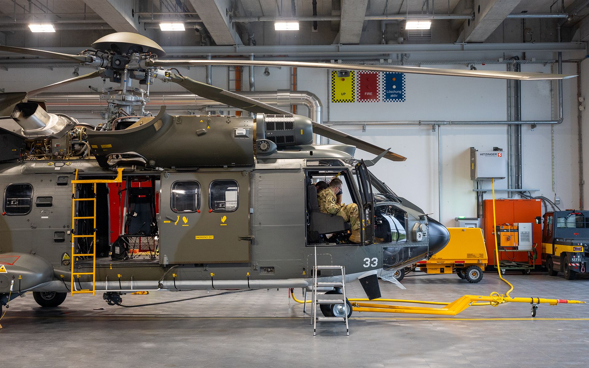 Ein militärischer Helikopter steht in einem Hangar. Eine Person sitzt vorne im Cockpit.