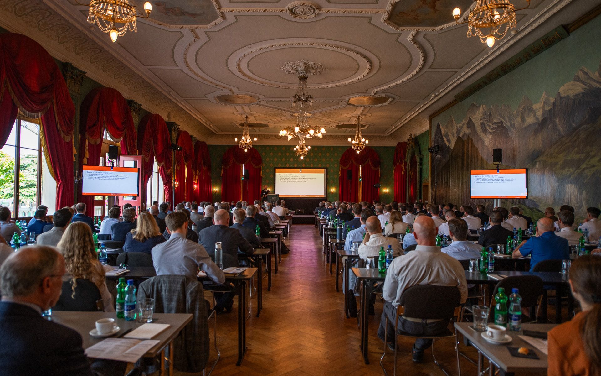 Viele Menschen sitzen an Tischen in einem grossen, barock-aussehenden Saal