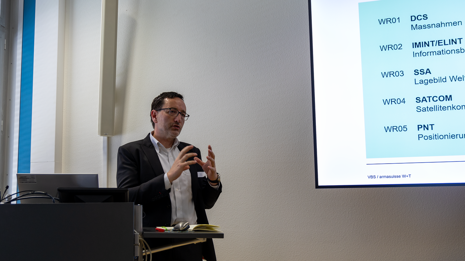 Dr Peter Erni stands behind a lectern. Next to this is a screen on which a presentation can be seen. 