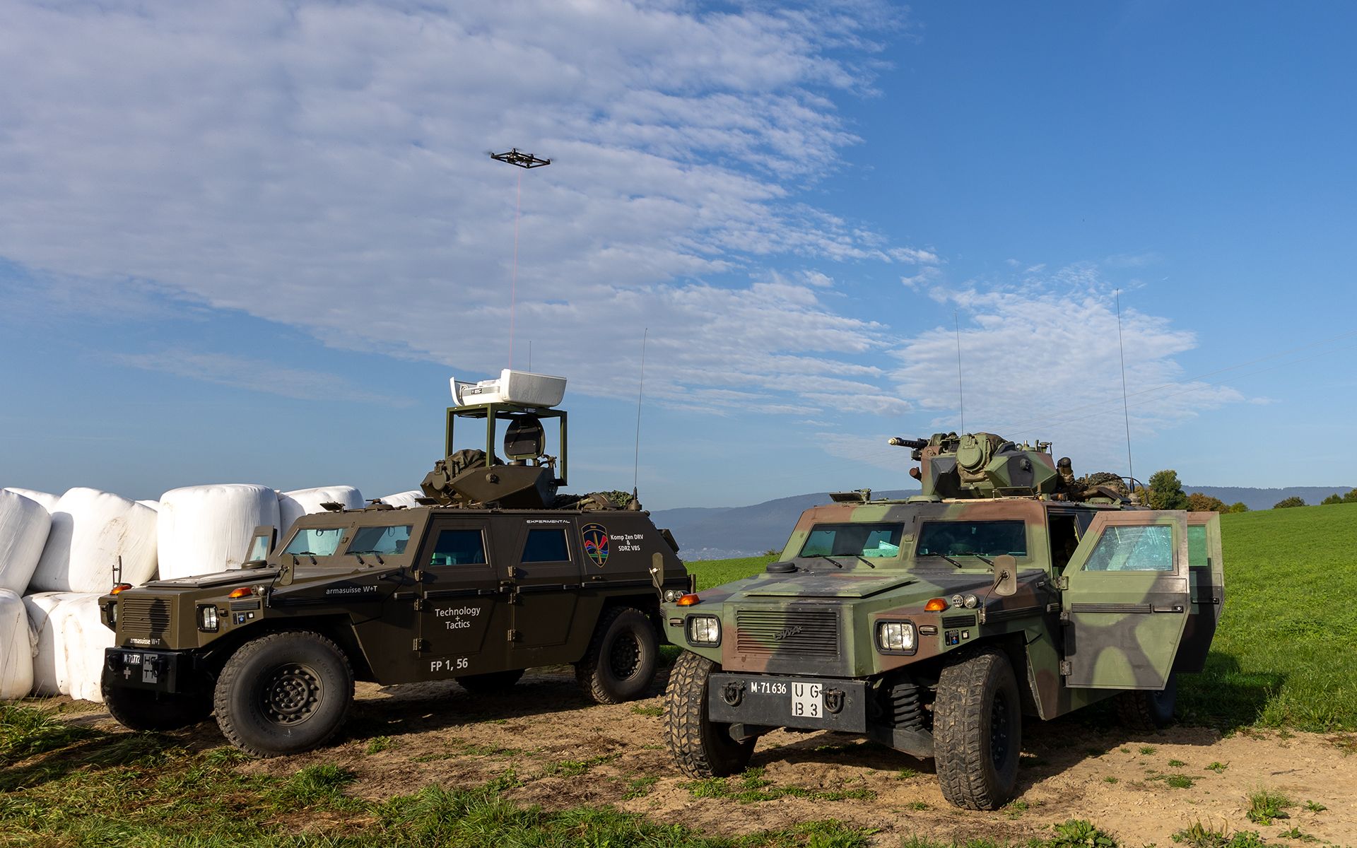 Two military vehicles are parked on farmland. A drone is flying in the background.