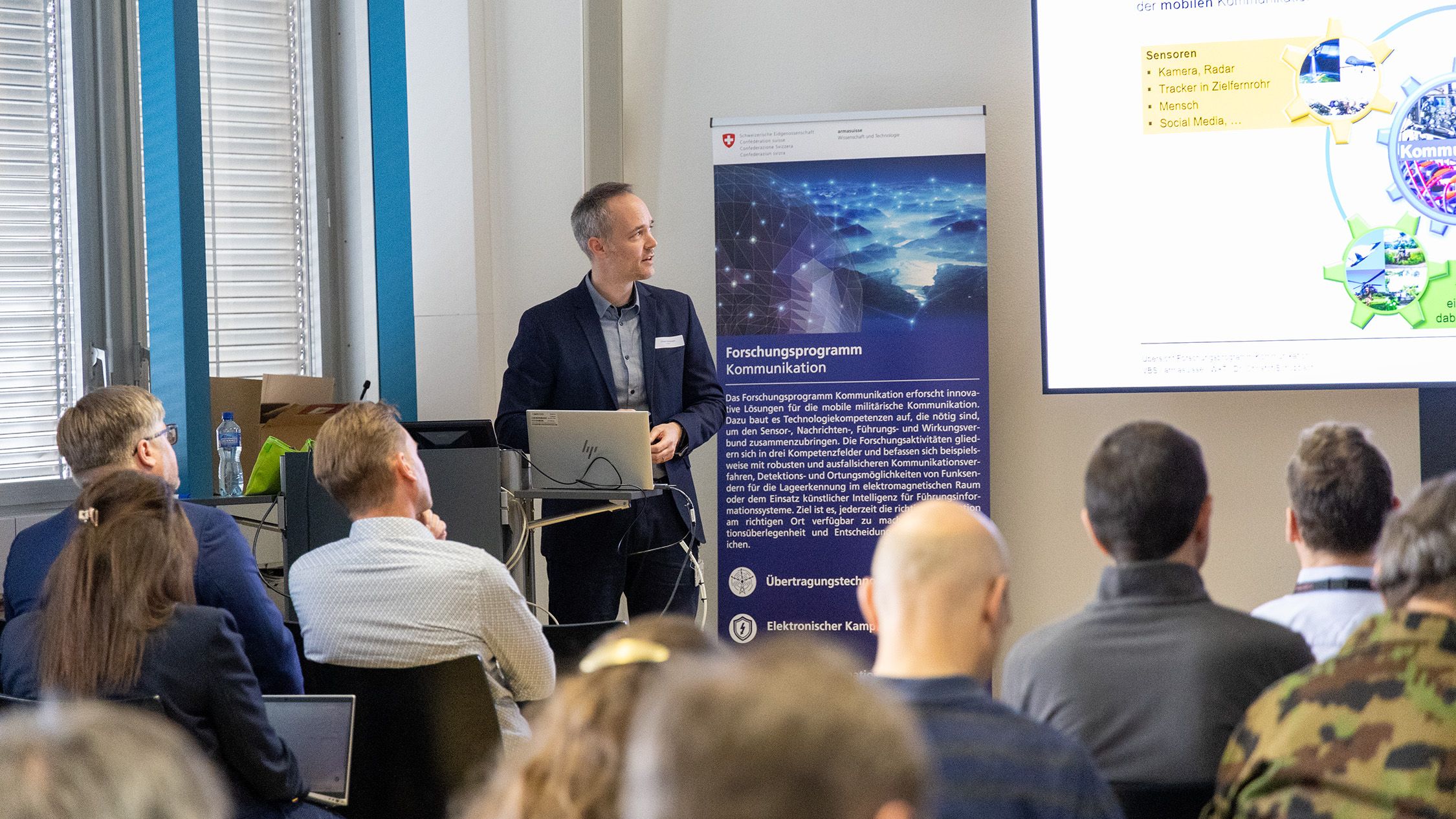 Dr Christof Schüpbach stands at the front of a room with chairs and speaks to an audience. The listeners are sitting in rows in front of him while he holds his presentation.