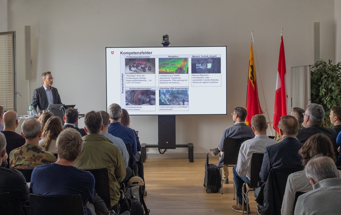 A man is standing in front of an audience and holding a presentation. A screen is positioned next to him.