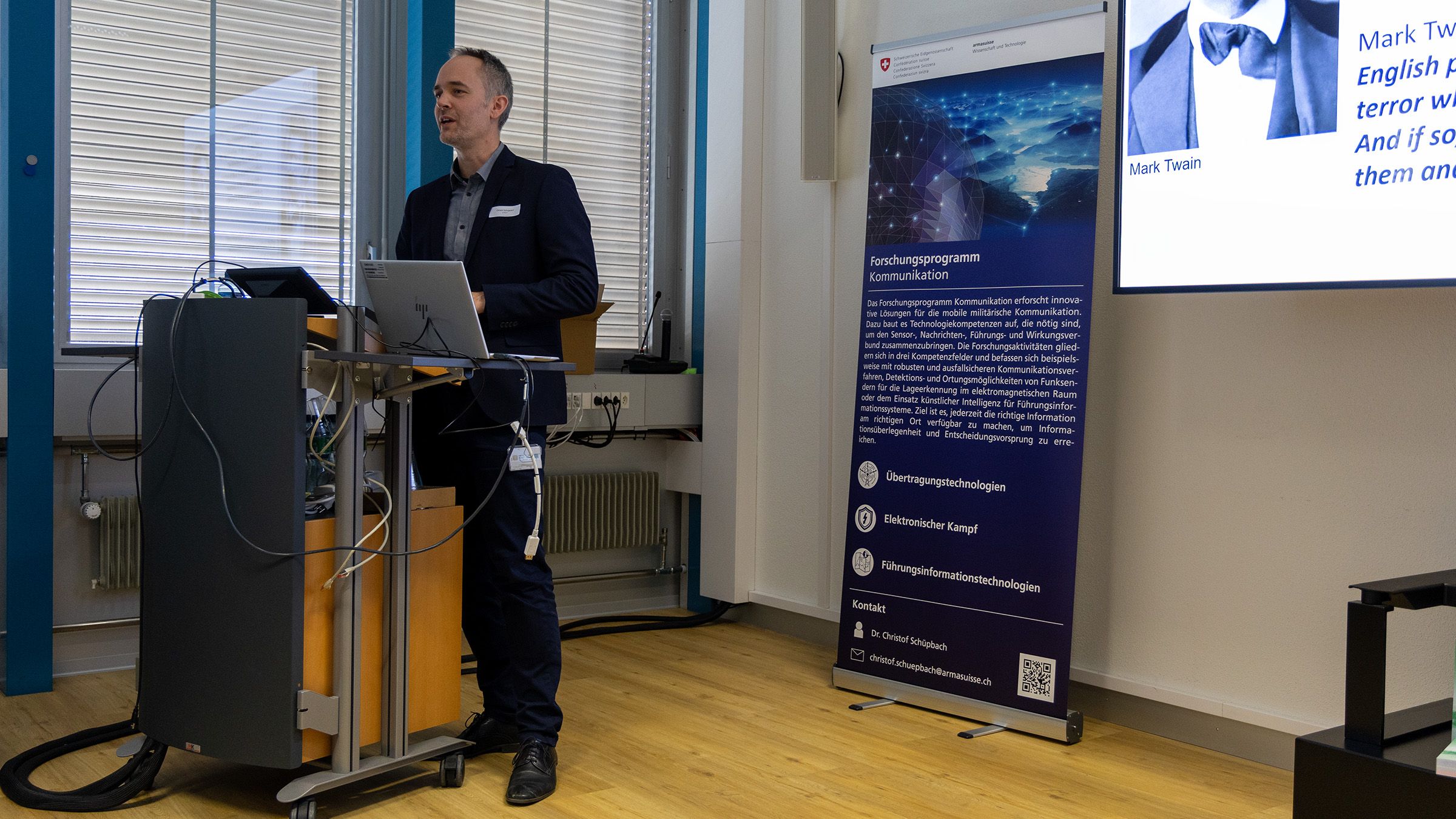 Dr Christof Schüpbach stands behind a speaker’s lectern next to a screen on which a presentation can be seen.