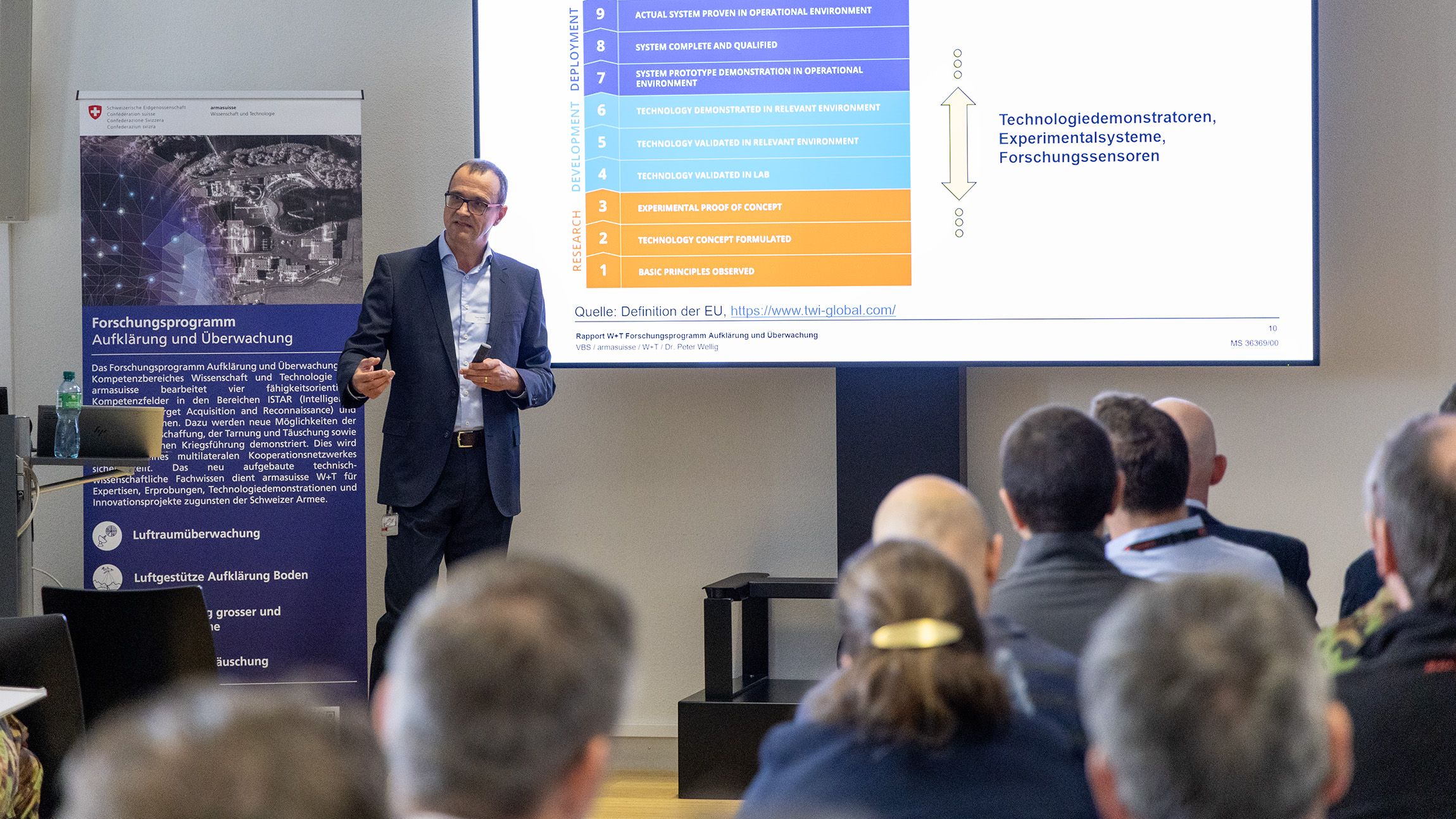 Dr Peter Wellig stands at the front of a room with chairs and speaks to an audience. The listeners are sitting in rows in front of him while he holds his presentation.
