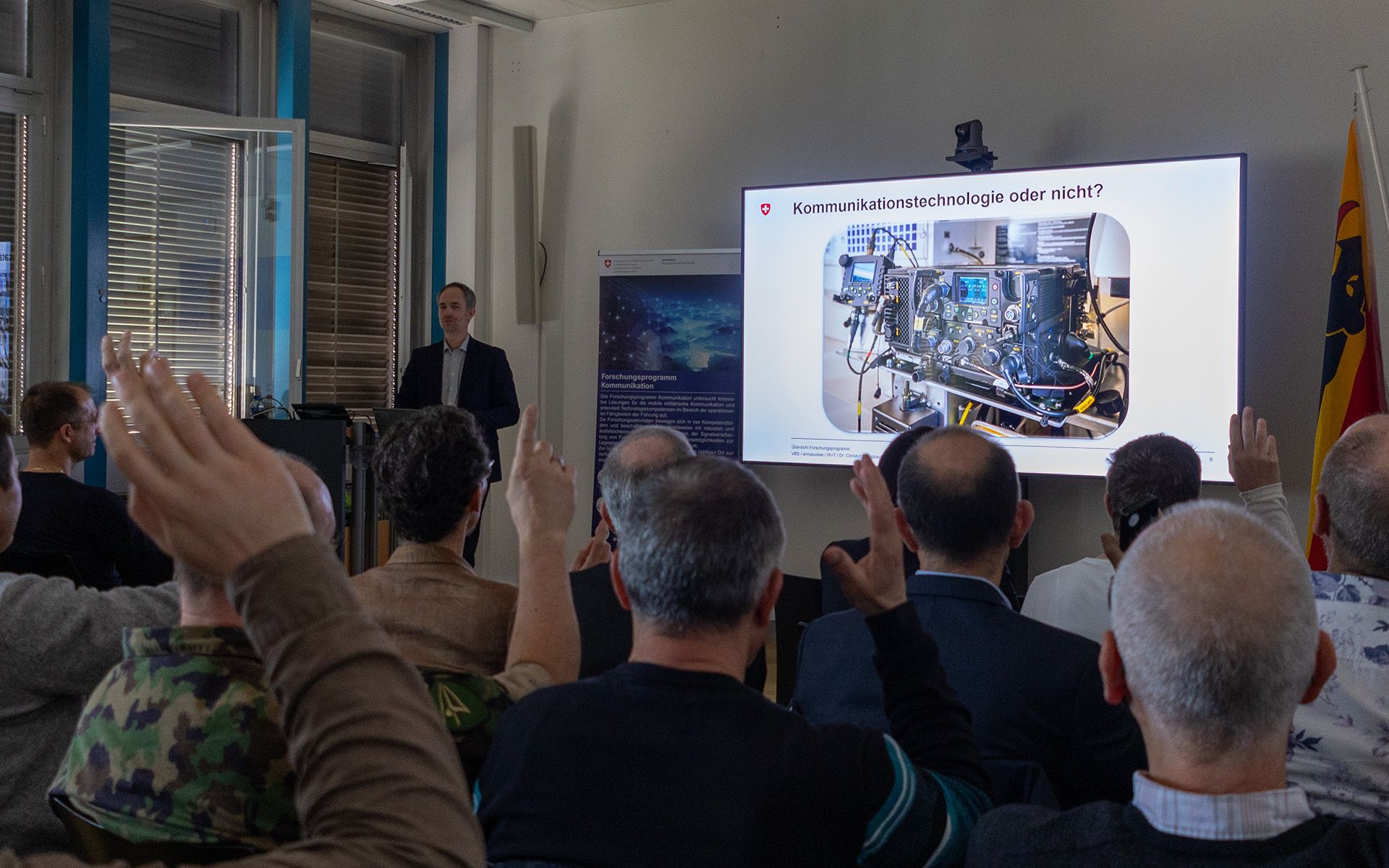 A man stands at a podium and presents a PowerPoint slide on a large screen. Facing him sits an audience, some of whom are raising their hands. 