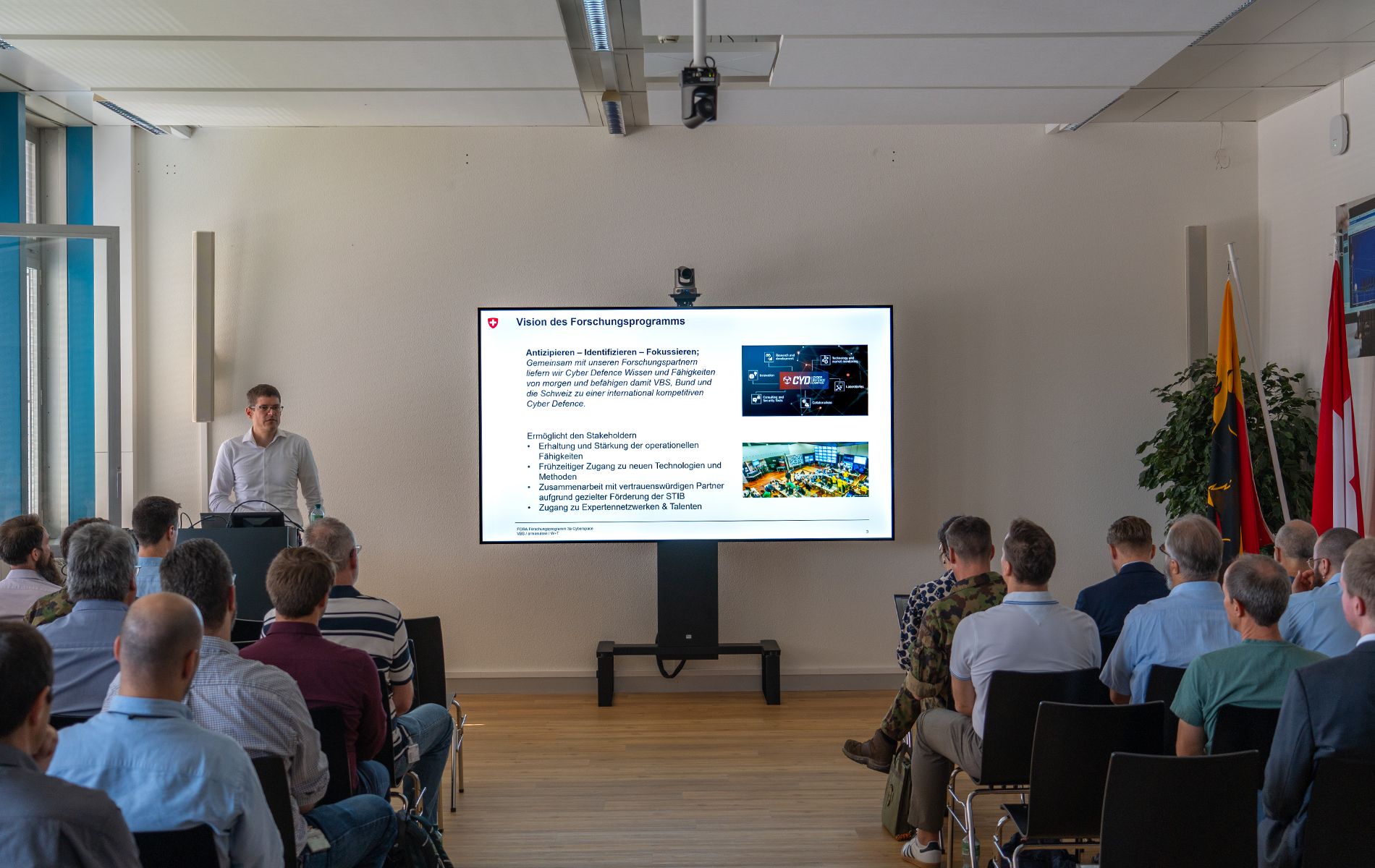 A man is standing next to a screen and holding a presentation. An audience is seated facing him.