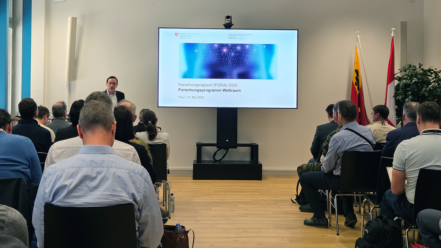 Dr Peter Erni stands at the front of a room with chairs and speaks to an audience. The listeners are sitting in rows in front of him while he holds his presentation.
