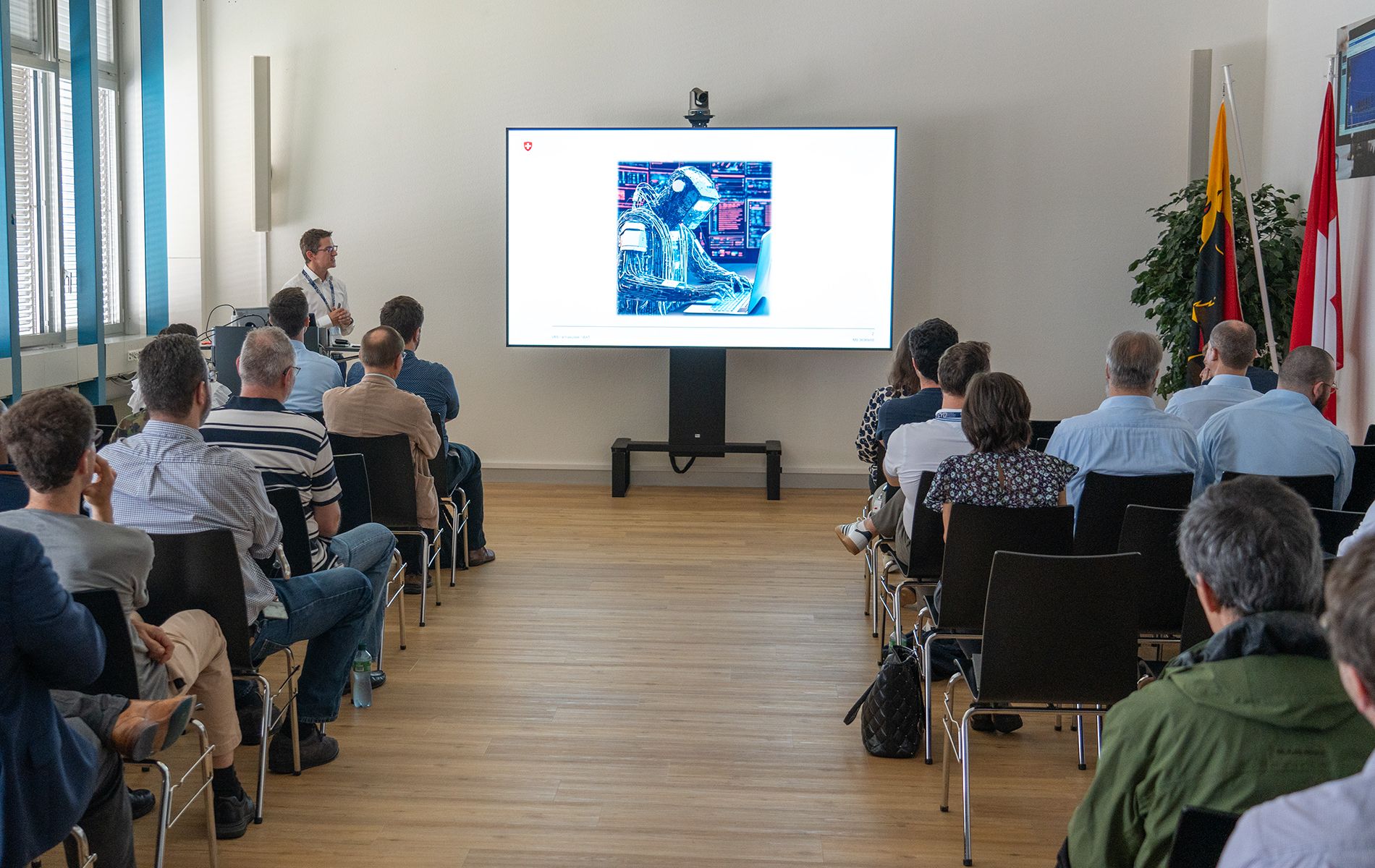 A man is standing next to a screen and holding a presentation. An audience is seated facing him. 
