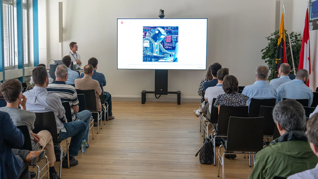 A man is standing next to a screen and holding a presentation. An audience is seated facing him.