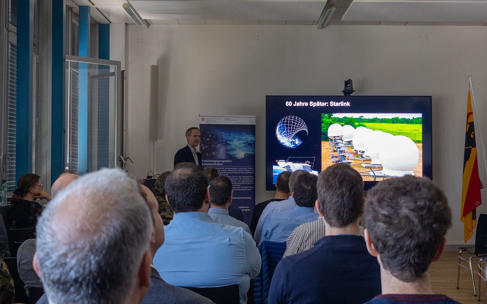 A man standing next to a large screen is holding a presentation. An audience is seated facing him. 