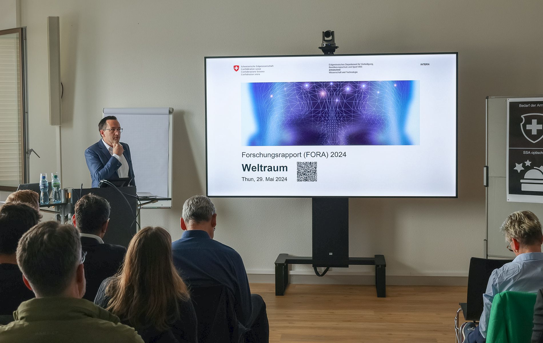 A man is standing in front of an audience and holding a presentation. Next to him is a projection screen.
