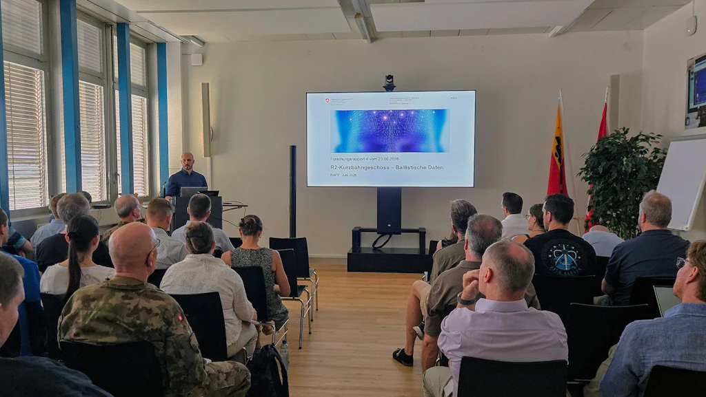 A man, standing next to a large screen, is giving a presentation. An audience is seated facing him.