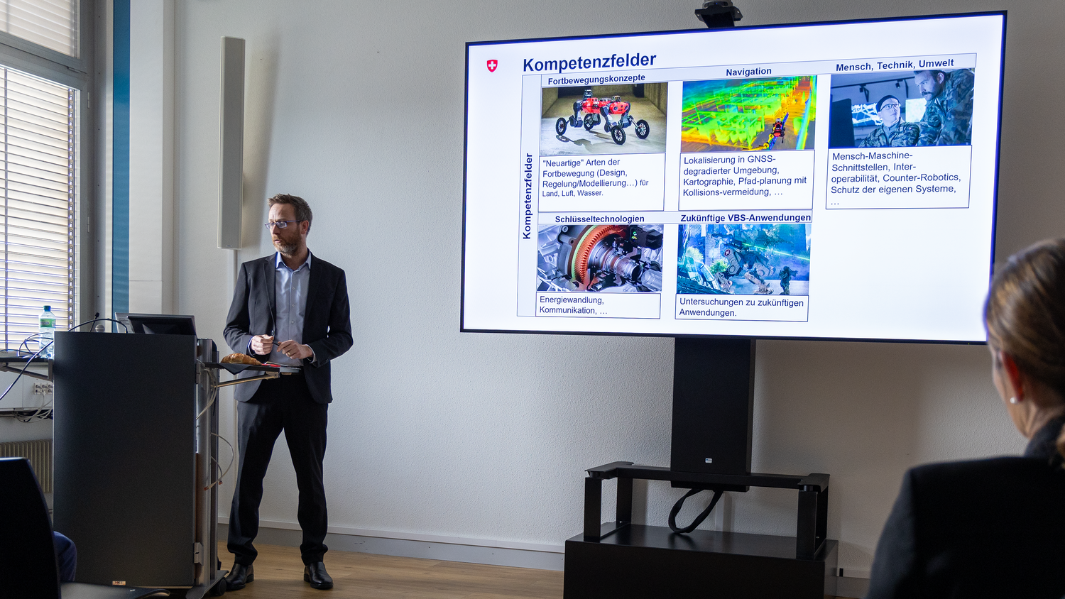 Dr Markus Höpflinger stands behind a lectern. Next to this is a screen on which a presentation can be seen.