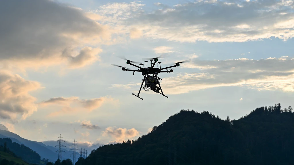 A drone flying in the sky. Clouds and landscape are visible in the background and it is getting dark.