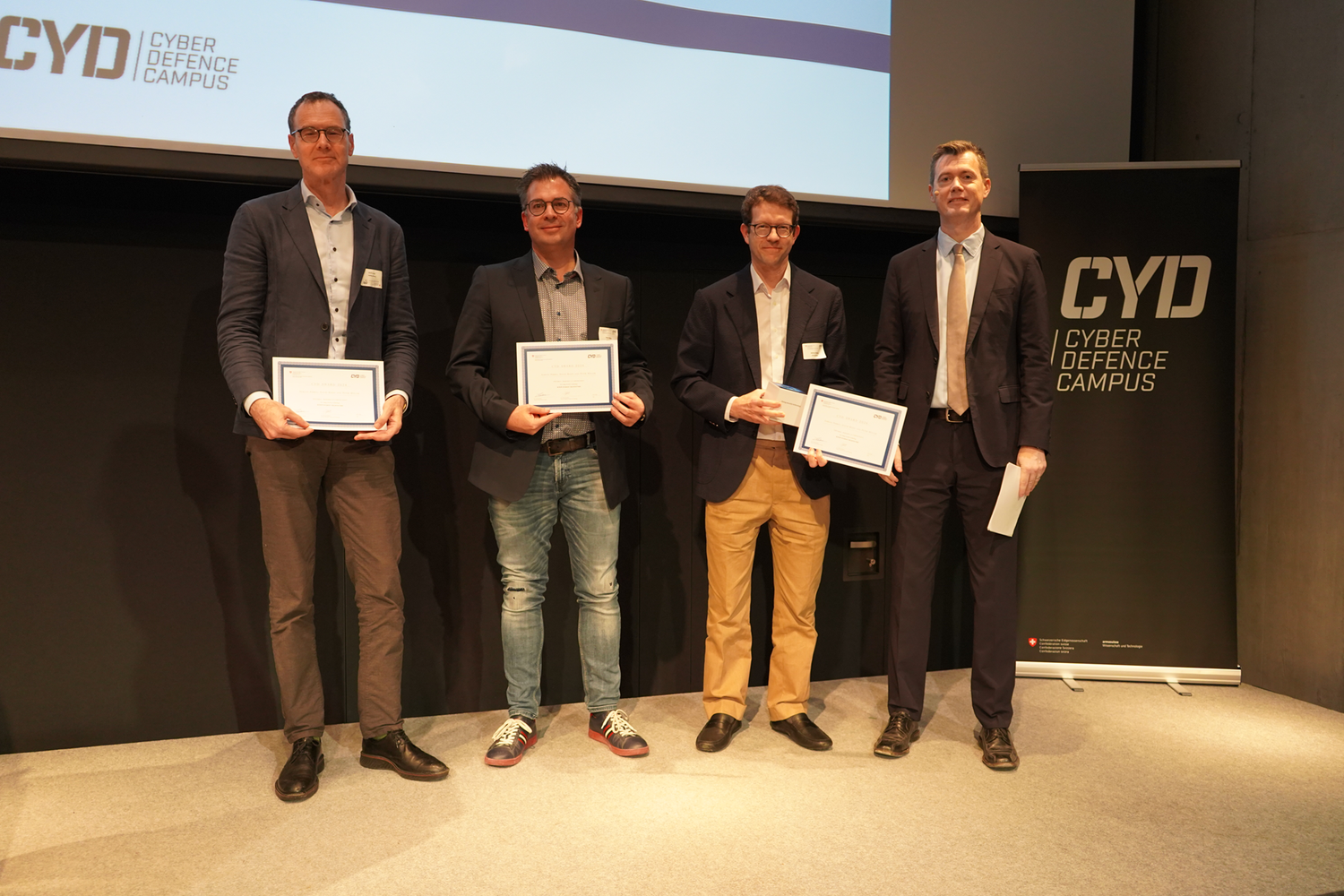 Quatre hommes se tiennent sur une scène, tenant des certificats dans leurs mains.