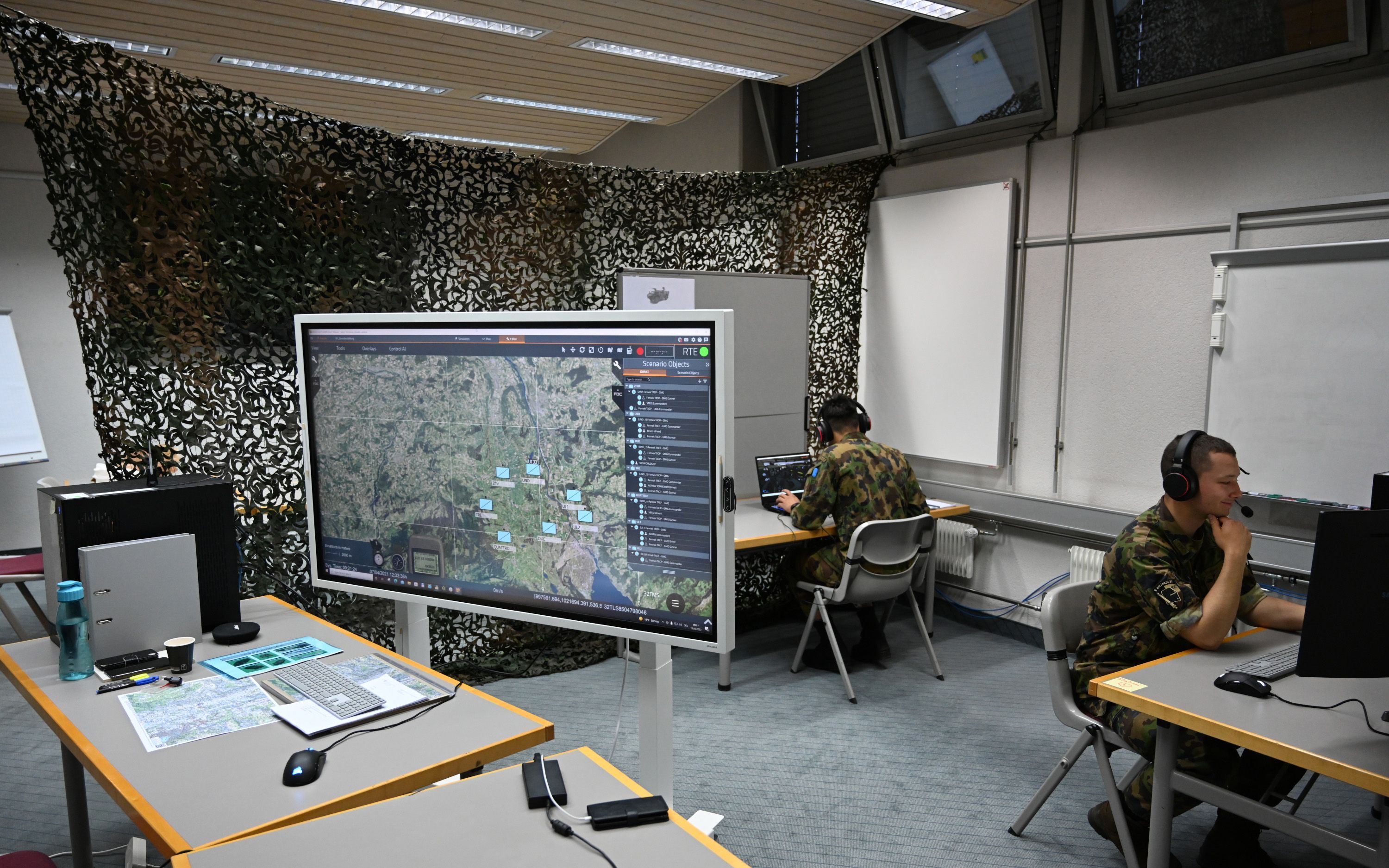Grand espace, qui est décoré à l’arrière avec un filet de camouflage vert. À droite de la photo, deux hommes en uniforme militaire sont assis à une table, dos à dos. Devant à gauche, on peut voir un écran géant et d’autres tables.