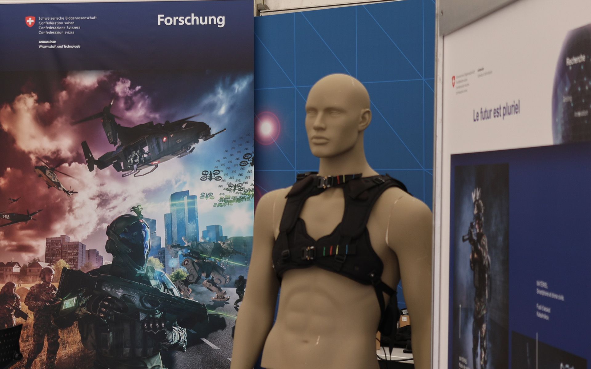 On voit une partie de l’espace d’exposition d’armasuisse S+T : des affiches, un mannequin, une table avec des brochures et l’arrière d’un écran. 