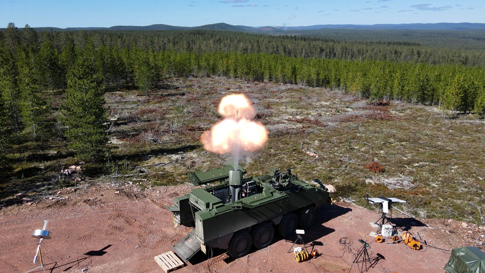 Au premier plan, l’observateur voit le système d’armes « mortier 12 cm 16 » lors d’un tir. Différents systèmes de mesure répartis autour du système d’arme sont visibles. 