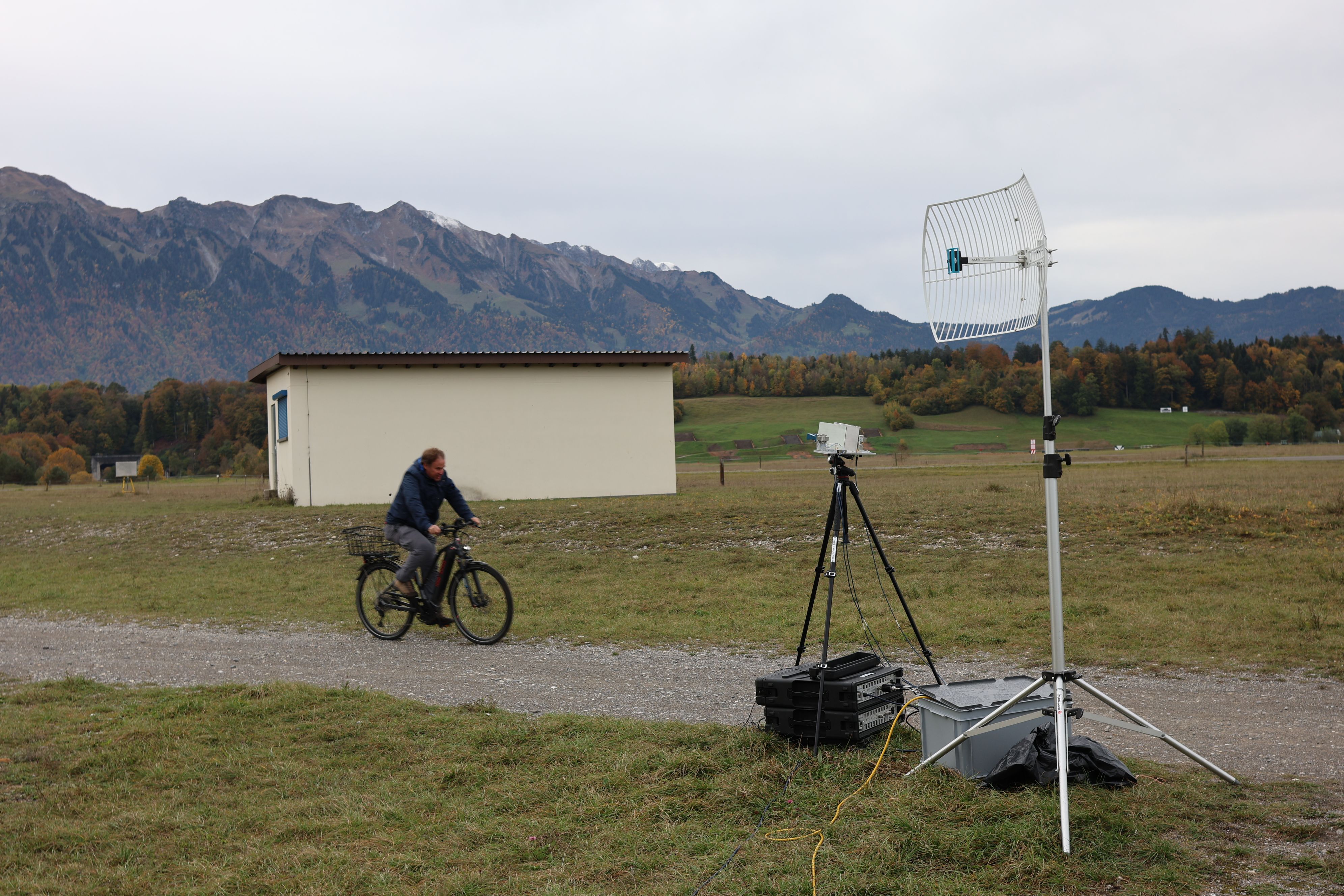 On voit au premier plan deux caisses, une antenne et un trépied noir. Un homme s’approche à vélo et on voit à l’arrière-plan une petite maison et un paysage. 