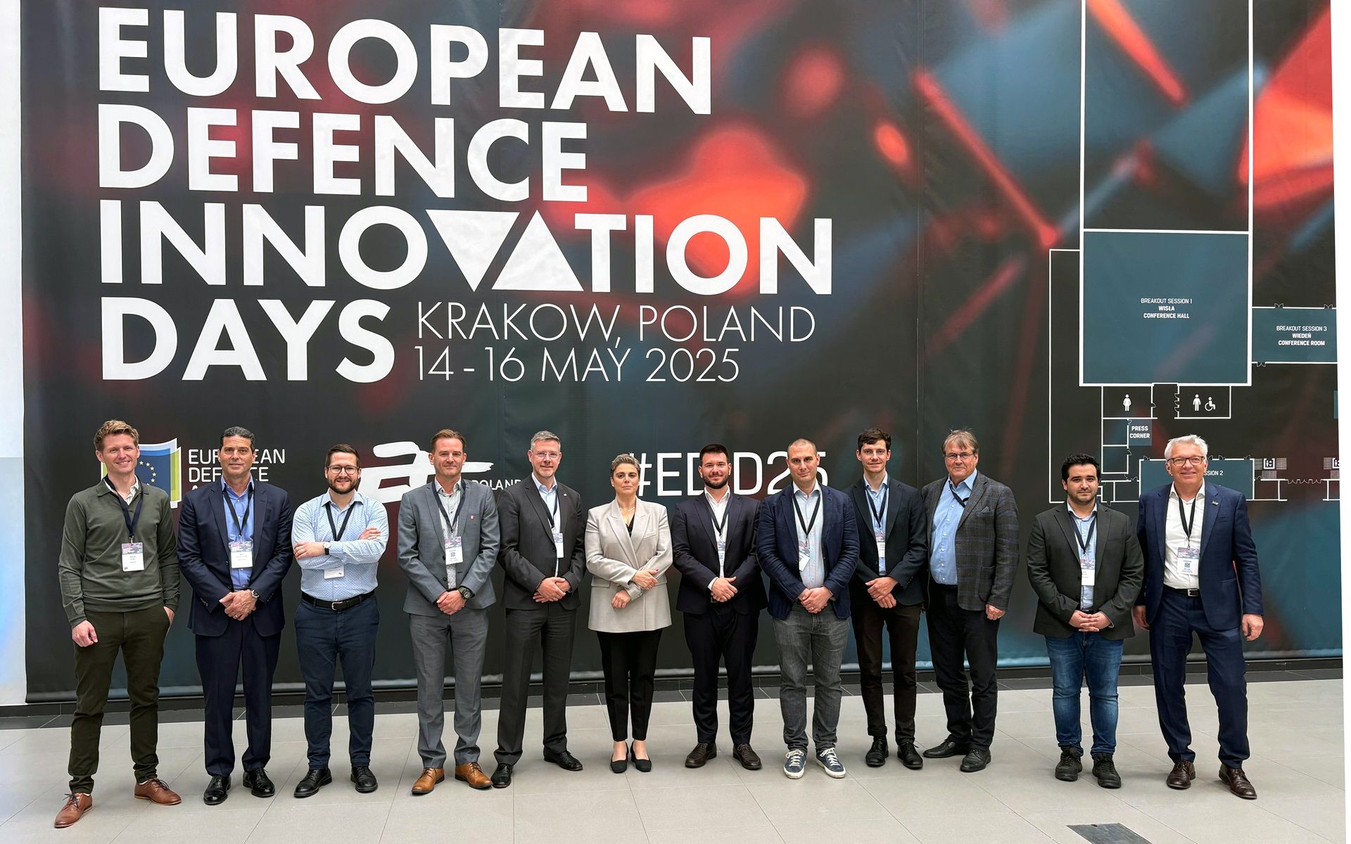 Des personnes se tiennent devant un mur sur lequel est écrit « European Defence Innovation Days, Krakow, Poland ».