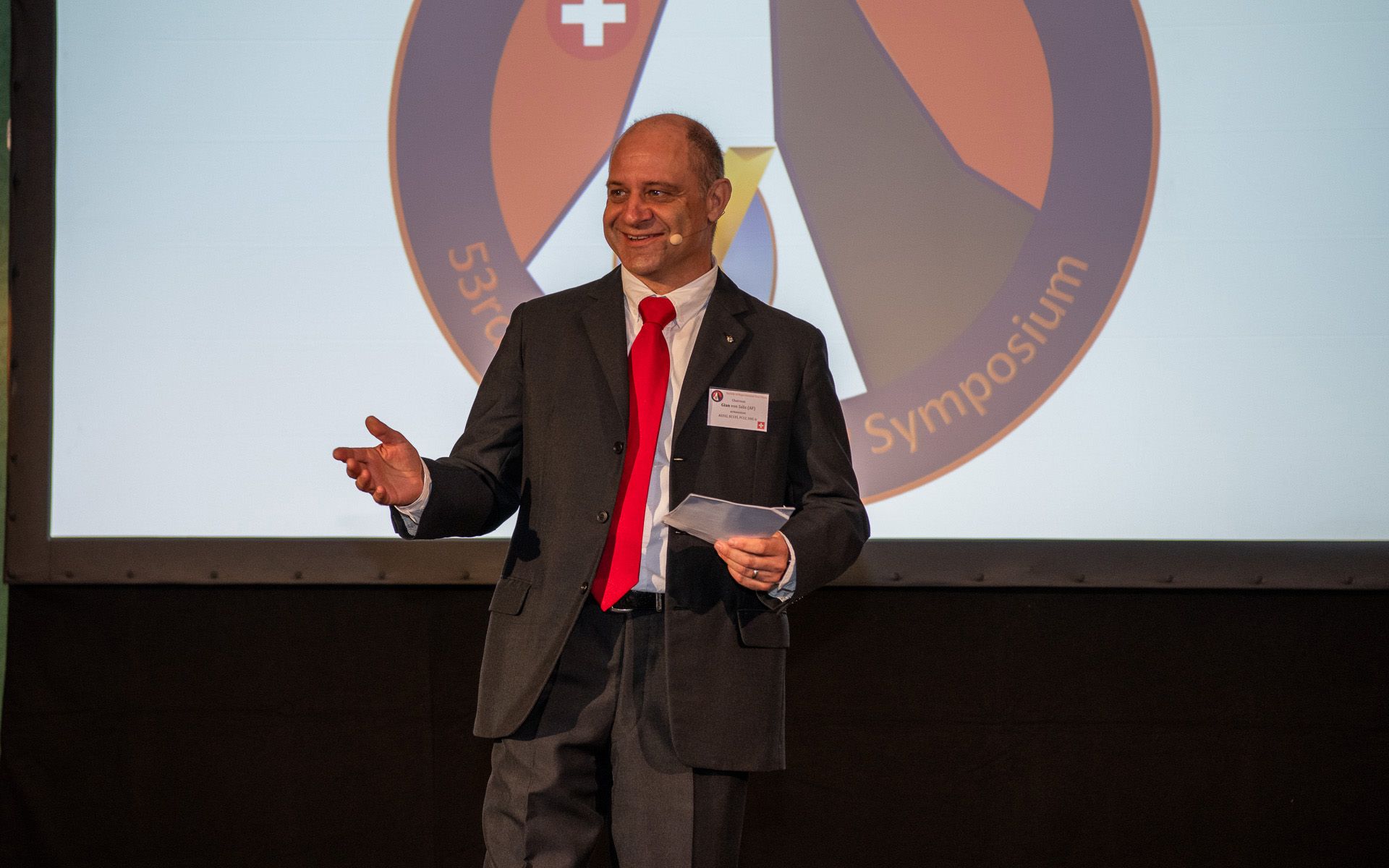 Un homme avec un costume noir et une cravate rouge se tient sur scène, avec une feuille dans la main.