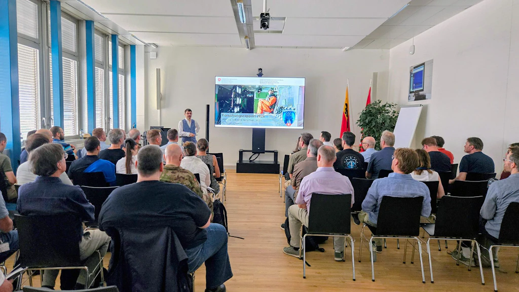Un uomo è in piedi in una sala, davanti a un leggio, e sta presentando una diapositiva in PowerPoint proiettata su un grande schermo. Di fronte a lui è seduto il pubblico.