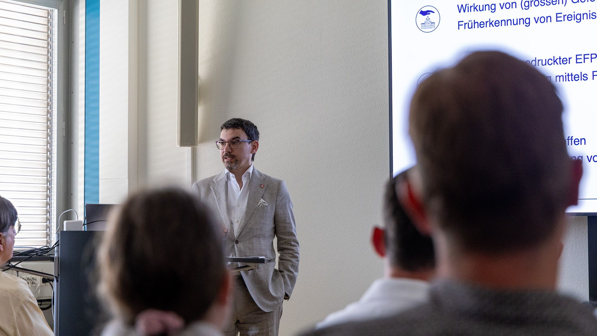 In piedi dietro a un leggio, con uno schermo al suo fianco, il dott. Ronny Lorenzo presenta al pubblico il programma di ricerca 4 «Efficacia bellica, protezione e sicurezza».