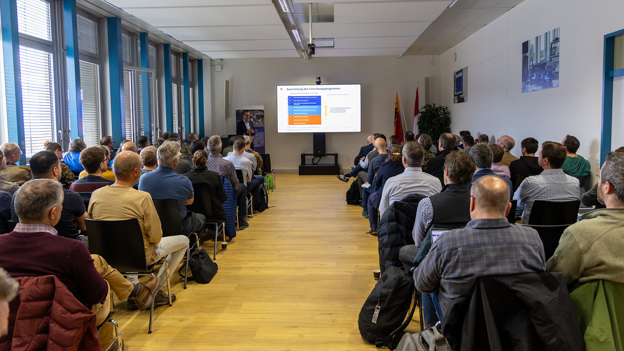 Il dott. Peter Wellig mentre parla al pubblico davanti a una sala gremita. Le persone del pubblico sono sedute su diverse file davanti a lui mentre tiene la sua presentazione.