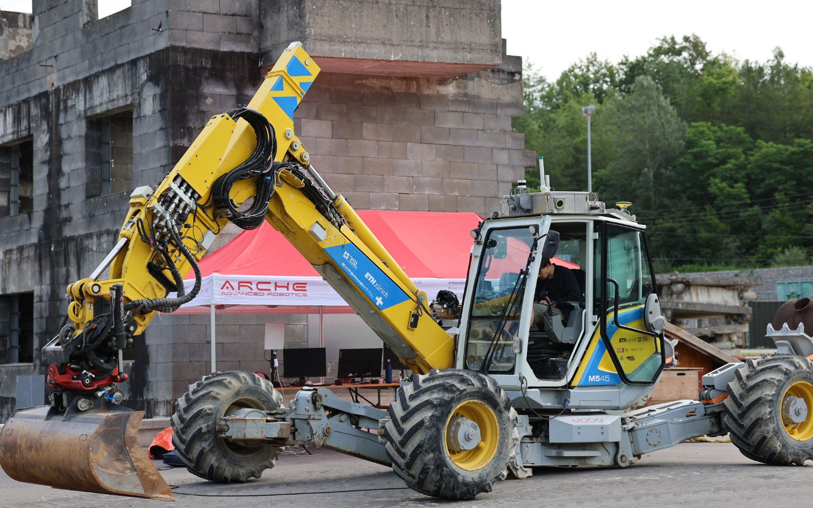 Un uomo è seduto dentro a un’escavatrice ragno gialla e, dietro a essa, si vede una tenda rossa con la scritta ARCHE. 
